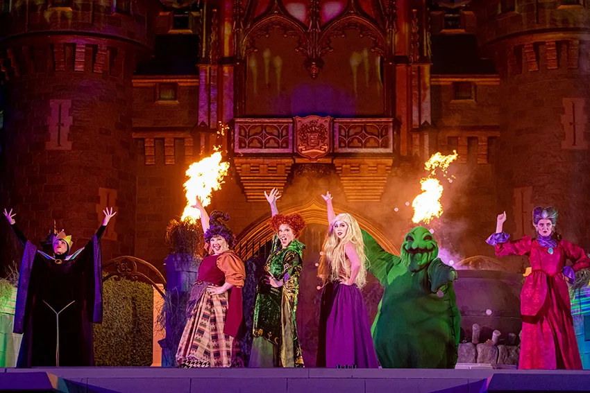 As irmãs Sanderson do filme Hocus Pocus no palco à frente do Castelo da Cinderela, cercadas por fumaça e efeitos de luz roxa, durante o show “Hocus Pocus Villain Spelltacular”