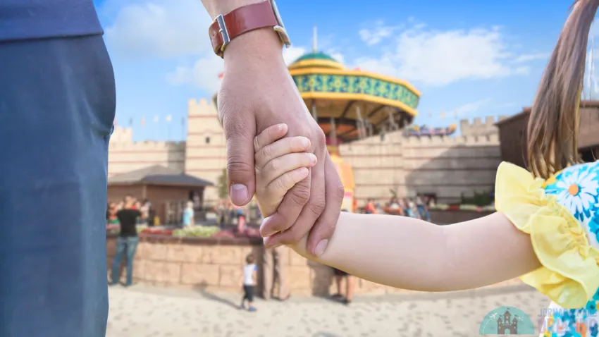 Uma criança está de mãos dadas a um adulto e, ao fundo, um parque de diversões